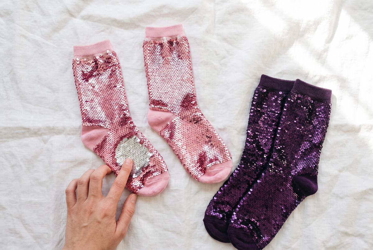 Main qui brosse les paillettes roses d'une chaussette posée à plat sur un drap blanc, à côté d'une paire de chaussettes à paillettes violettes