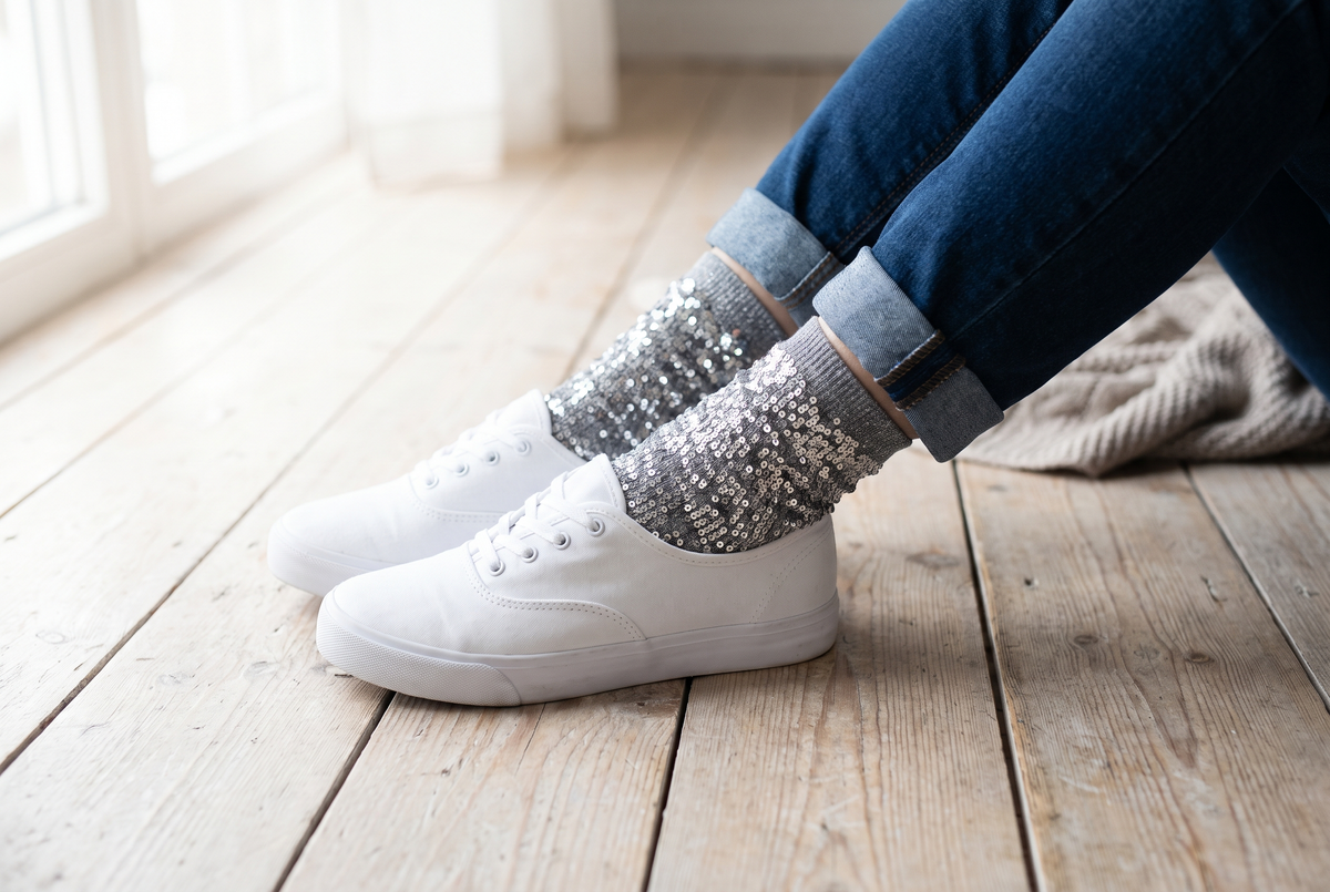 Pieds croisés portant des chaussettes à paillettes grises aux reflets argentés, avec un jean retroussé et des baskets blanches sur un parquet clair Image