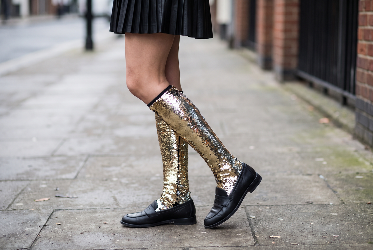 Jambes vues de côté portant des chaussettes hautes à paillettes dorées sous une jupe plissée noire, avec des mocassins noirs Image